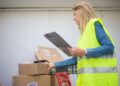 Woman in high-visibility vest handling boxes for delivery service inside a warehouse setting.